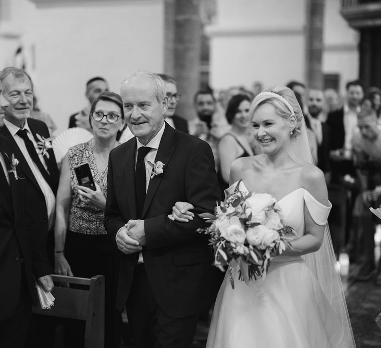 Bride wears pearl bridal headband as she carries white rose bouquet and walks down the aisle with her father