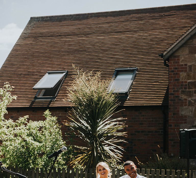 Bride in off the shoulder wedding dress sits holding hands with the groom for their outdoor humanist wedding ceremony 