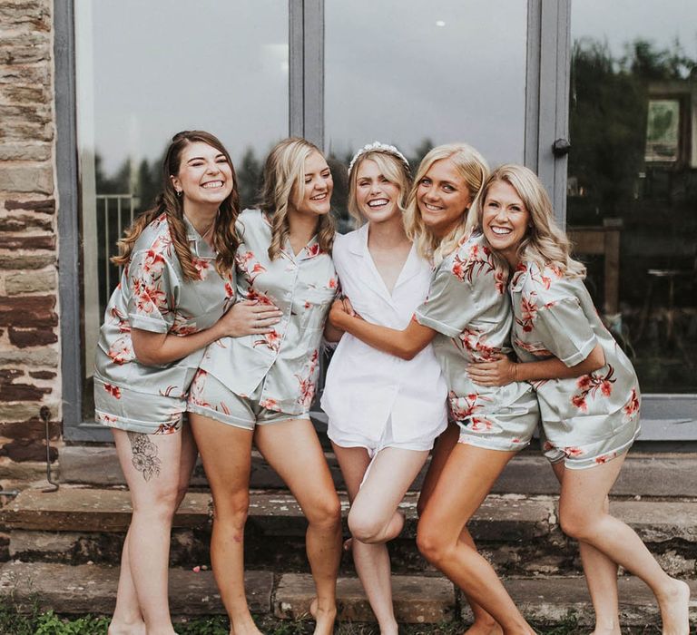 Bride in a white pyjamas and headband stands with the bridesmaids in green and pink flower pyjamas