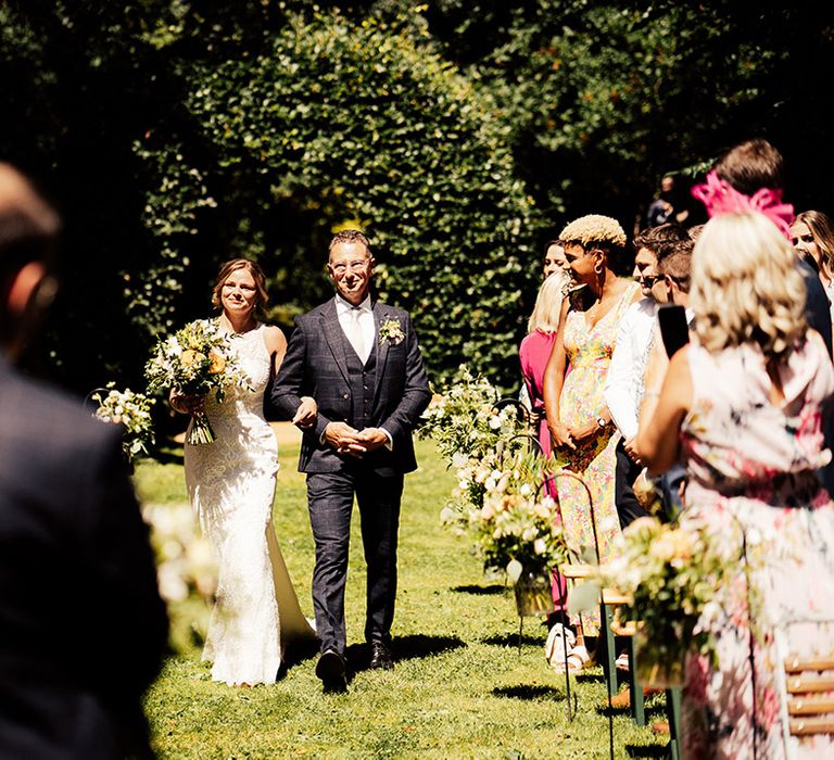 Father of the bride in a checkered blue suit walks with the bride in a fitted lace wedding dress down the aisle for outdoor wedding 