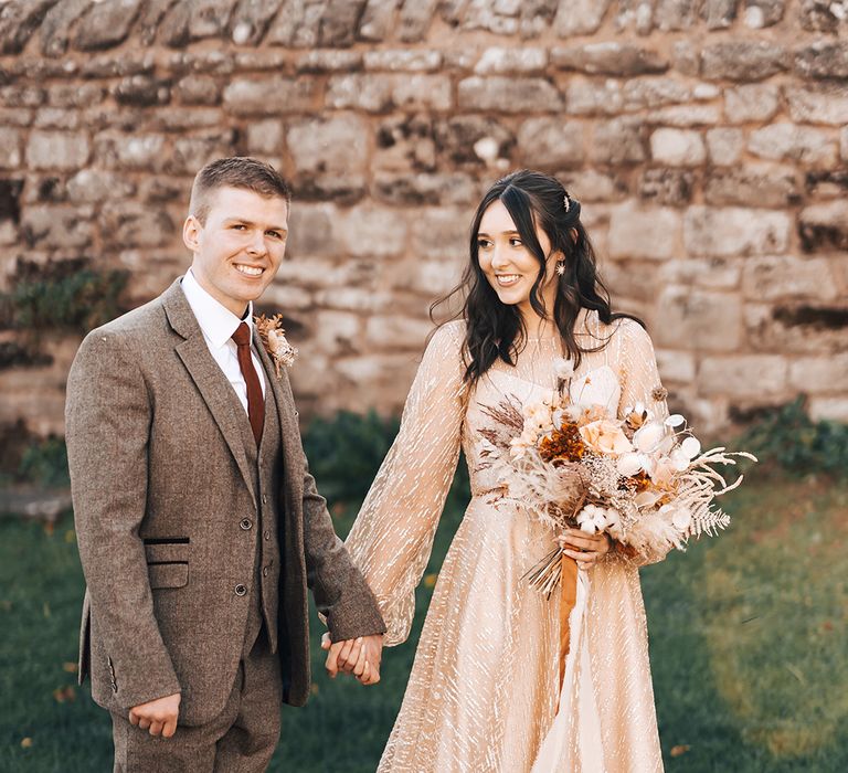 Bride in sheer gold wedding dress holds dried floral bouquet tied with burnt orange ribbon and walks with her groom in brown suit