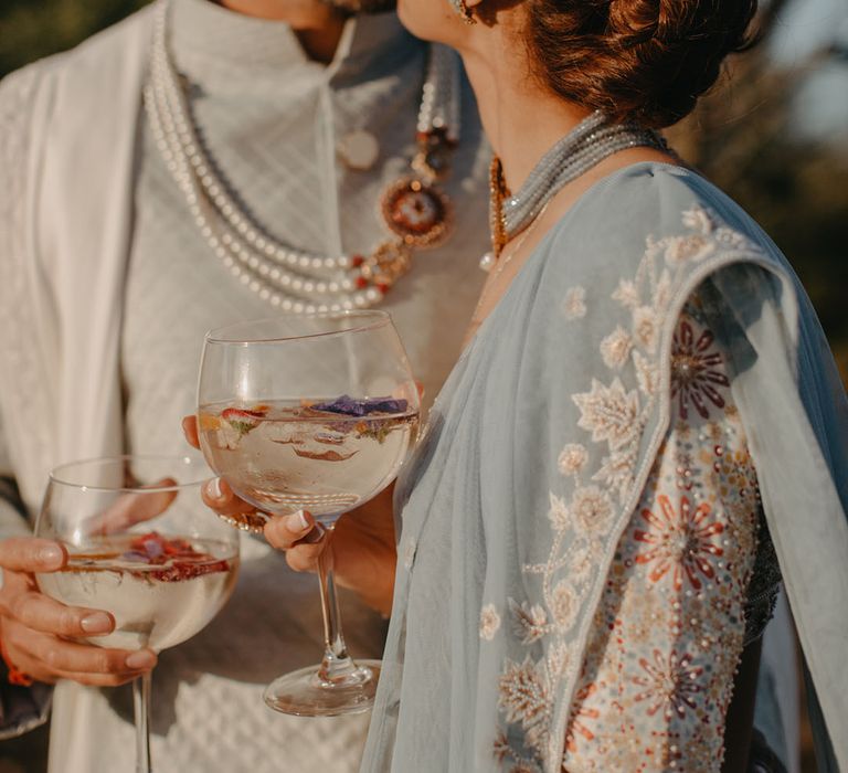 Bride wears her hair in intricate plaited style and colourful bridal outfit whilst stood with her groom during golden hour