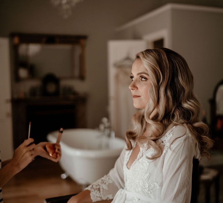 Bride in a white robe with lace details sitting in a chair getting her makeup done for her wedding 