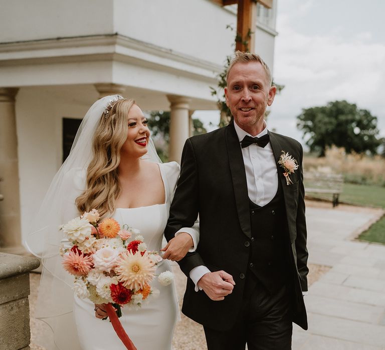 Bride in square neck long sleeve satin wedding dress with two tier veil holding colourful mixed bouquet with wedding guest 