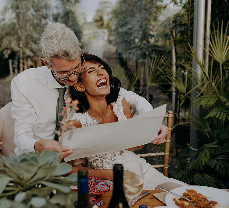 Laughing Bride leans on Groom who is reading while seated on rustic chairs