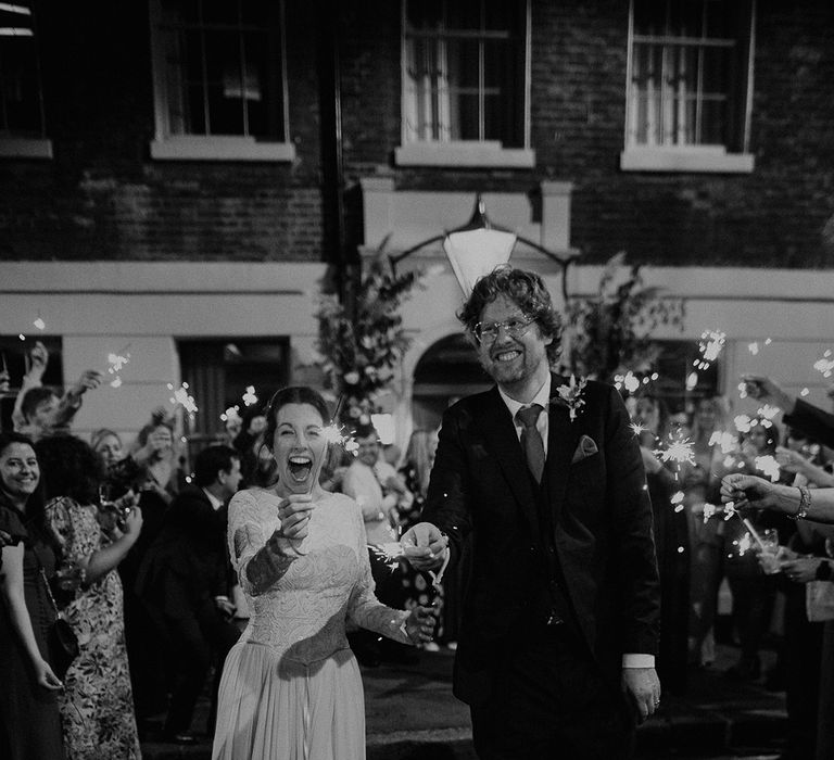 Groom in dark suit with orange tie and autumnal boutonniere and bride in long sleeve lace vintage wedding dress and closed toe satin heels doing sparkler exit from autumnal intimate London wedding