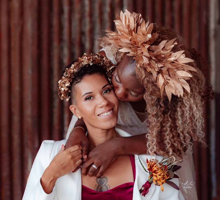 Bride in white silk suit with autumnal dried floral boutonniere and golden bridal crown being hugged by bride in large golden leaf bridal crown 