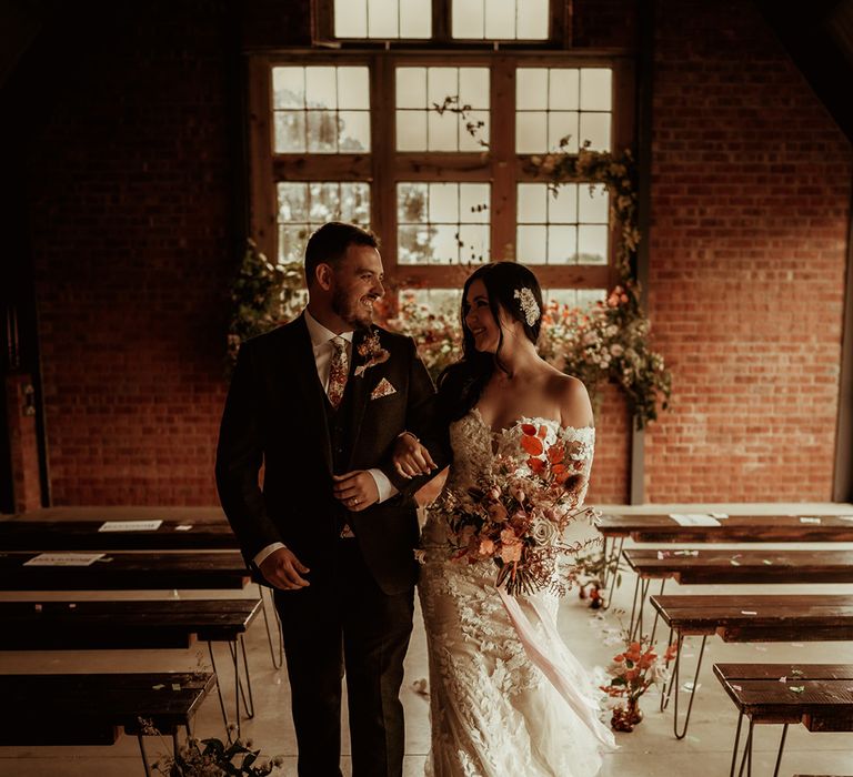 Groom in a dark suit with flower patterned tie and pocket square and dried flower boutonniere and bride in  off the shoulder bodycon wedding dress with long, sheer sleeves and lace detailing with a dried flower bouquet with roses, eucalyptus, peonies and autumn leaves linking arms and walking back down the aisle