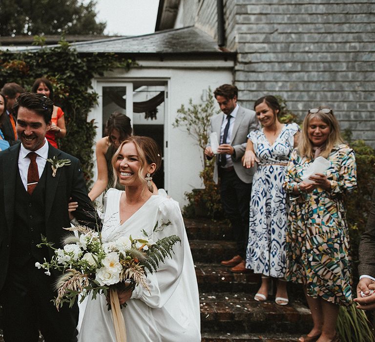 Bride holds white floral bouquet tied with ribbon after intimate wedding ceremony and walks alongside her groom in three piece suit