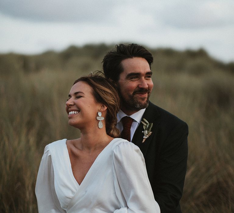 Bride in silk wrap wedding dress and groom in three piece suit embrace outdoors beside the sea