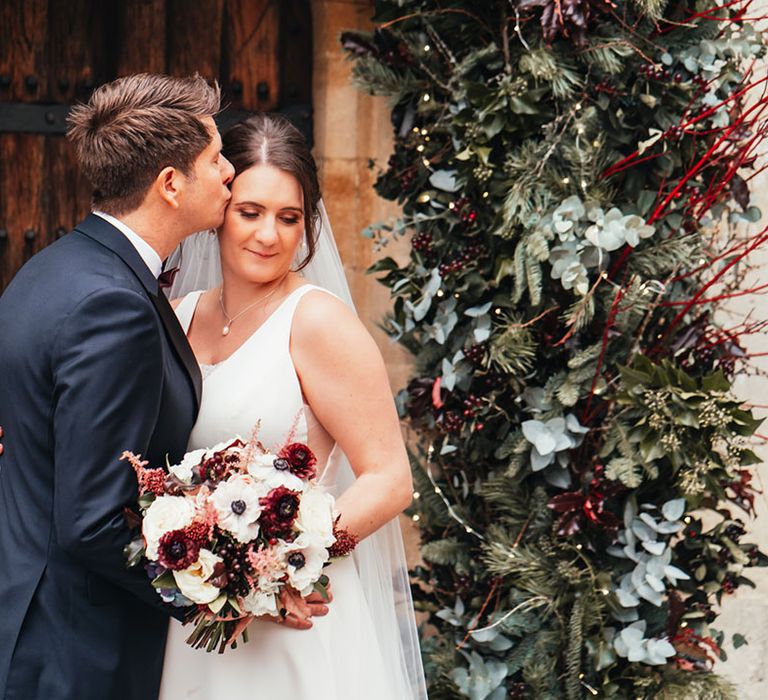 Groom in black tie kisses the bride on the forehead as she closes her eyes holding a white anemone and red flower bouquet 