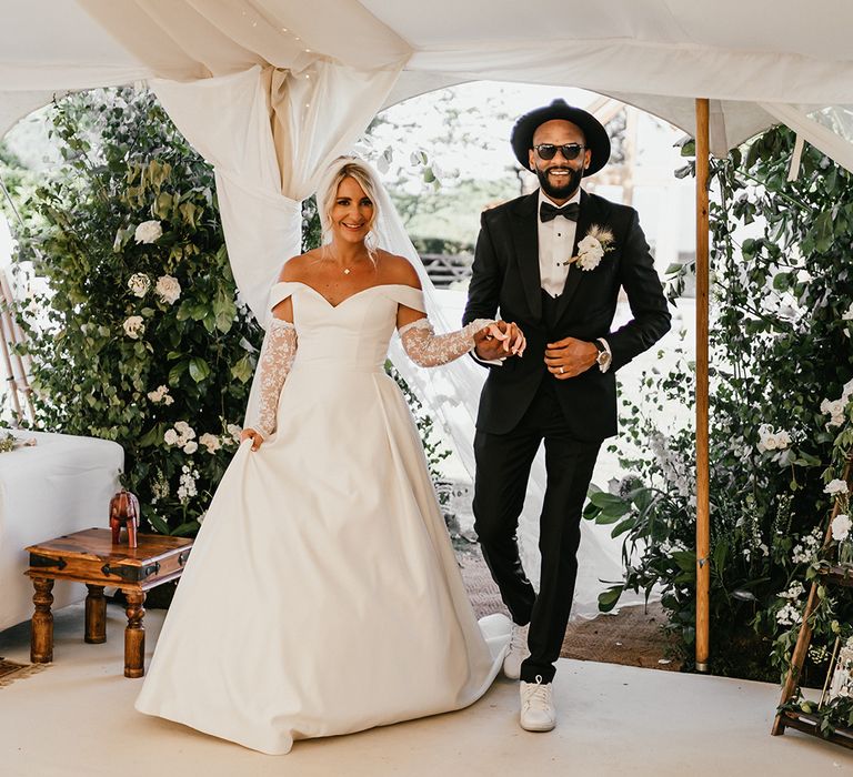 Blonde bride in off-the-shoulder princess wedding dress and lace sleeves walks alongside her groom in fedora and three piece suit