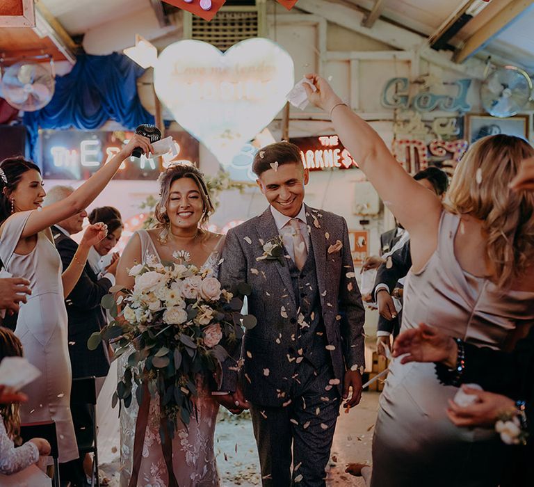 Bride holding large white rose, eucalyptus and foliage bouquet and groom in three piece grey suit walking down the aisle of Gods Own Junkyard doing confetti throw 