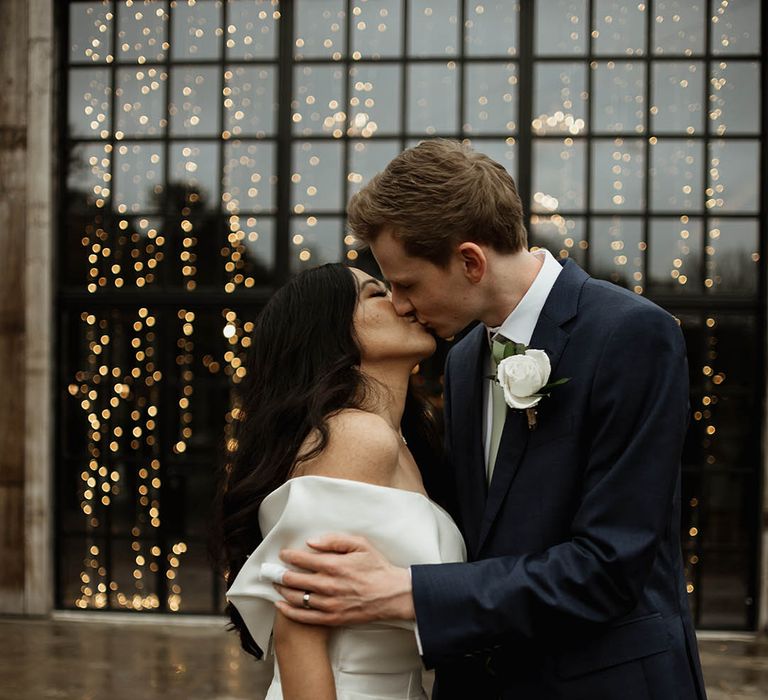 The bride in a classic off the shoulder gown kisses the groom in a navy suit and white rose buttonhole 