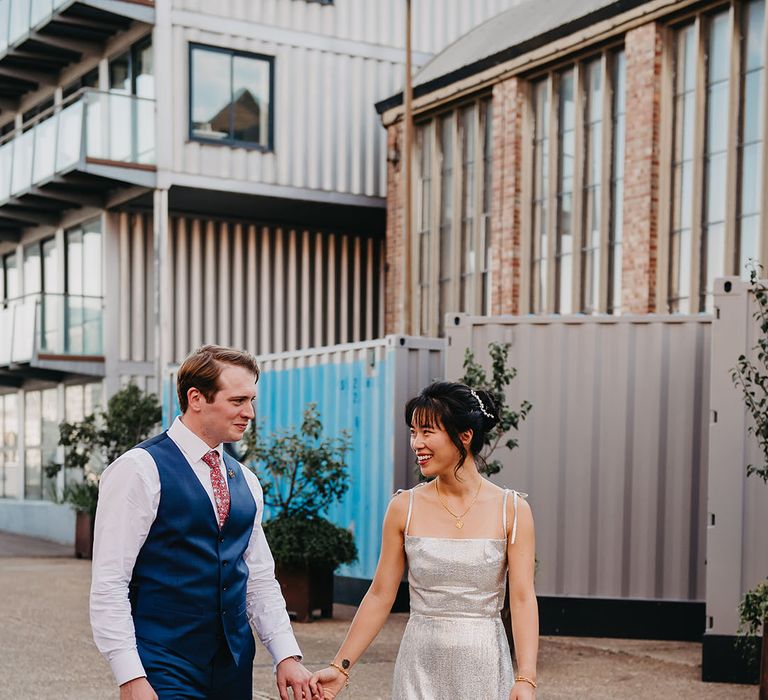 Bride in silver wedding dress walks alongside her groom in white shirt and blue waistcoat 