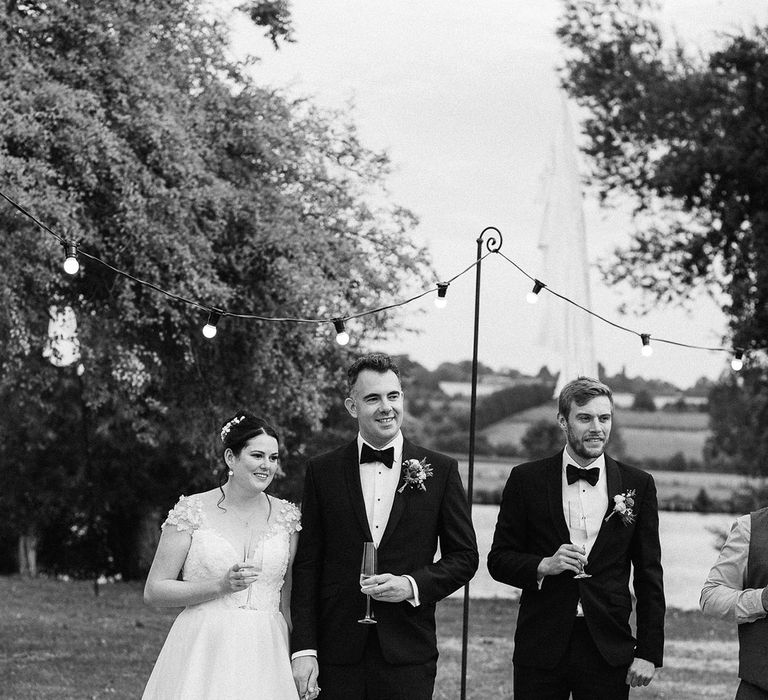 Festoon lighting decorates the outdoor area with the bride and groom standing together in their timeless wedding outfits sharing some wine 