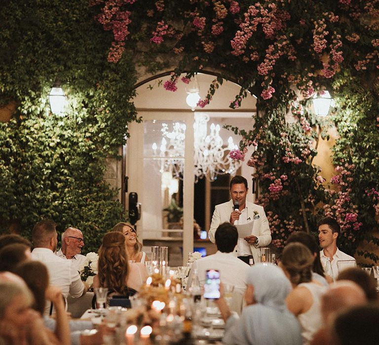 Groom gives outdoor speech beside beautiful green foliage and pink florals across Villa Di Ulignano