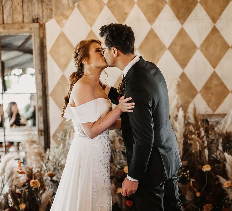 Bride in off-the-shoulder Love Spell wedding dress kisses her groom during barn wedding with Autumnal styling 