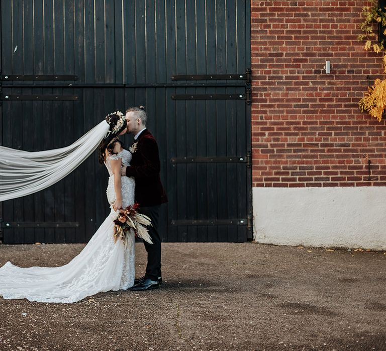 The groom in a burgundy velvet suit jacket kisses the bride in an off the shoulder lace wedding dress with the veil blowing in the wind 