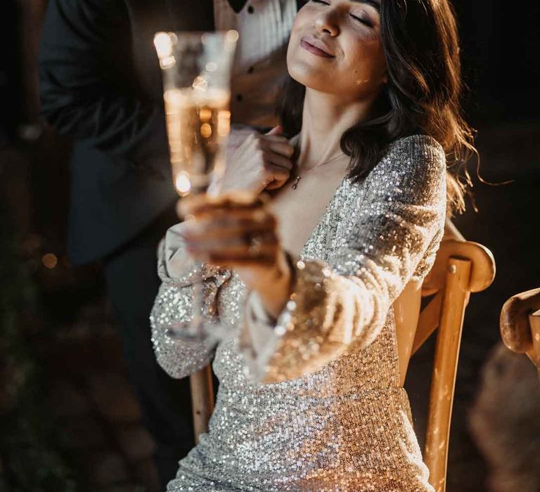 Bride in silver sparkly wedding dress holding glass of champagne, being kissed on the forehead by groom in classic black tuxedo and black bowtie 