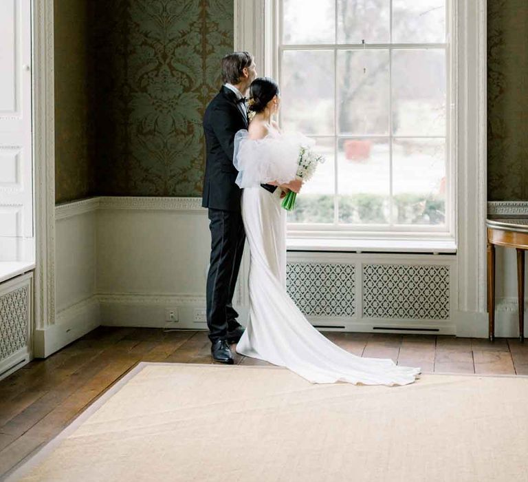 Bride in strapless wedding dress with puff tulle sleeves and groom in classic black tuxedo, black bowtie and boutonniere embracing at St Giles House