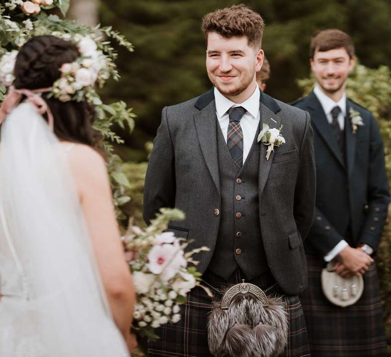 Groom wearing dark grey blazer and waistcoat, dark tartan tie with white flower boutonniere, dark grey and blue tartan kilt and wedding sporran looking at bride 