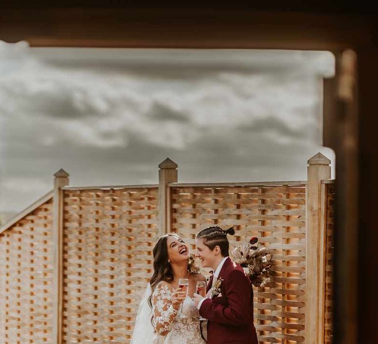 Bride in long sleeve lace illusion sleeve wedding dress with overlay drinking champagne with bride in maroon bridal suit with grey waistcoat, dusky pink tie and autumnal dried flower boutonniere 