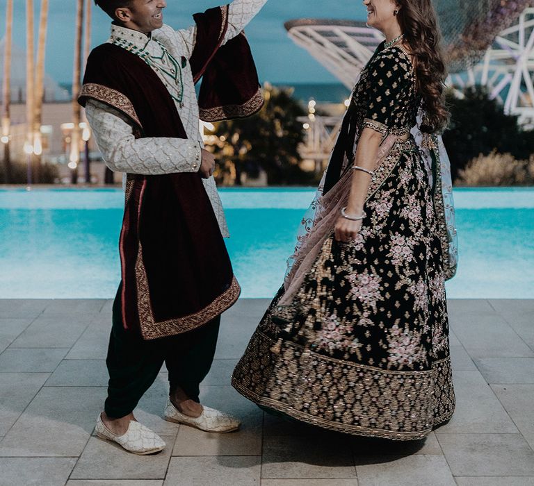 Bride in two piece black and gold patterned saree dancing with groom in white and gold patterned sherwani by the pool at castle wedding venue in Barcelona 