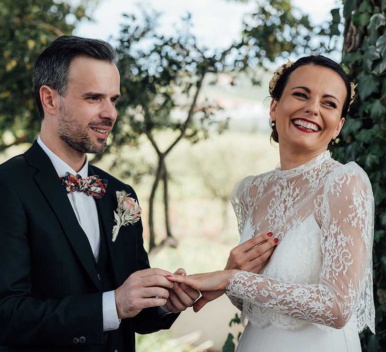 bride in a lace wedding dress with long sleeves and groom in a black suit and bow tie exchanging wedding rings