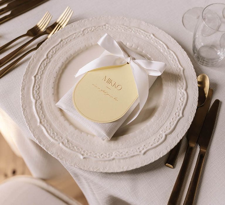 Pastel yellow and white wedding table setting with white napkin, yellow place name and white bow with gold cutlery 