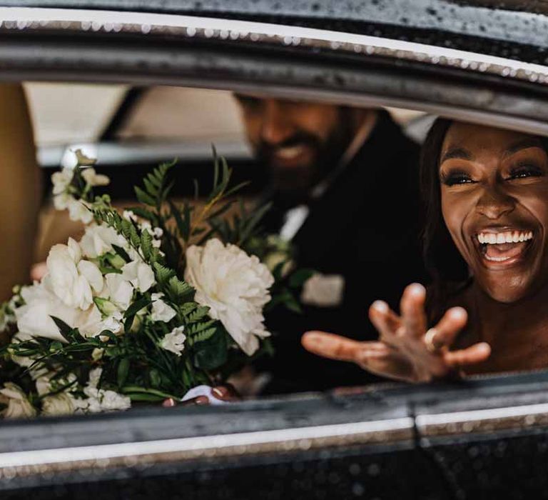 Bride holding white garden rose, white hydrangea, cows parsley and foliage bridal bouquet waving at guests from classic black wedding car 