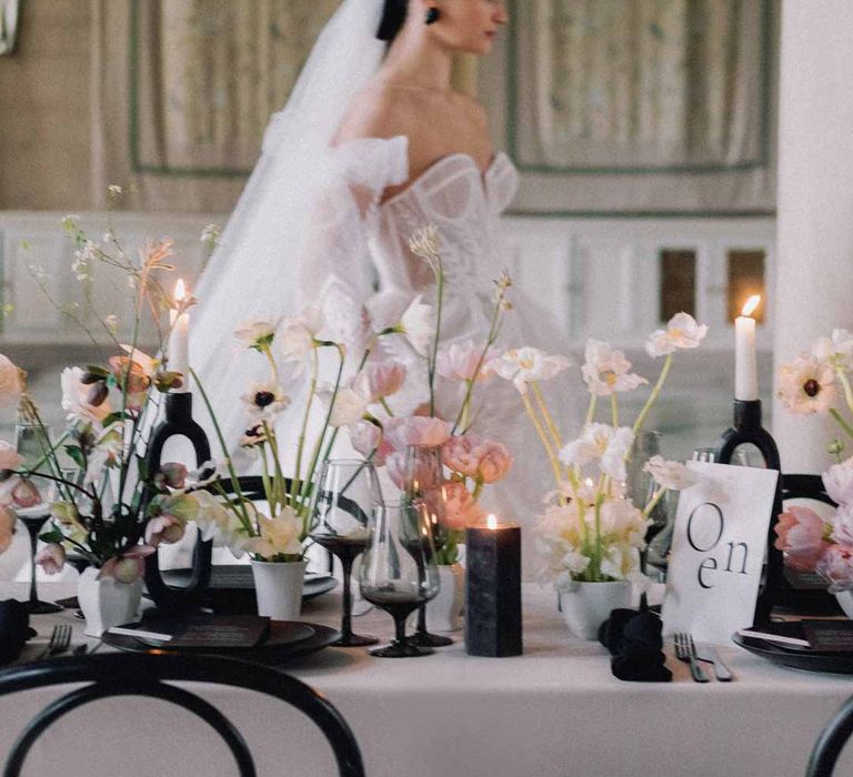 Bride wearing sweetheart neckline tulle mesh bridal gown with detachable puff tulle sleeves and delicate beading with cathedral length veil walking by Notan inspired wedding tablescape consisting of light pink wedding tablecloth, white poppies, light pink Persian buttercups, baby's-breath and foliage centrepieces, black artistic candlestick holders, white tapered candles, black crockery and monochrome wedding stationery 