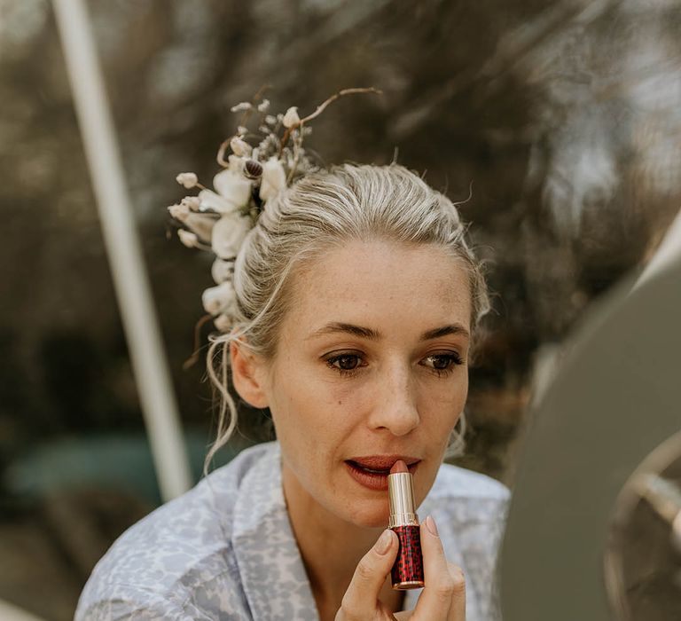 Bride in light blue and white wedding pyjamas doing her own wedding makeup with her hair in a sleek updo with white flower hair accessory 