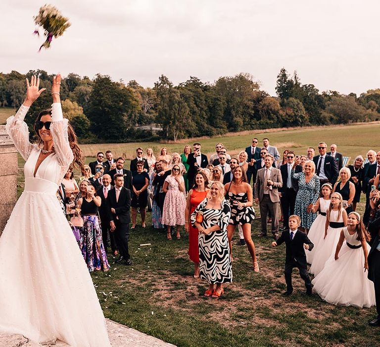 The bride in a pearl long sleeve wedding dress tossing her bouquet the wedding guests hoping for a proposal