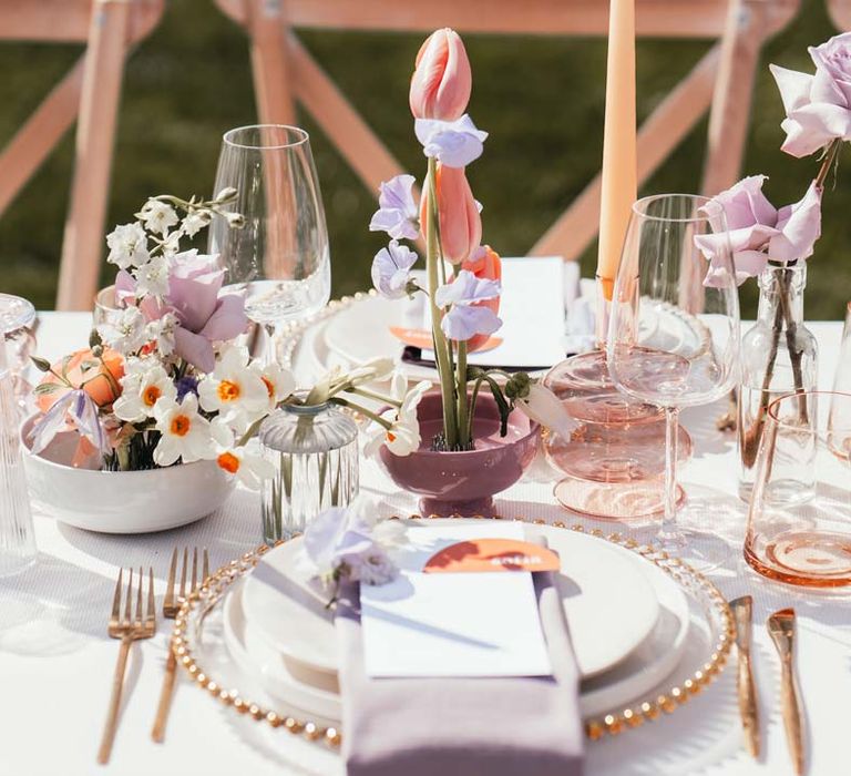 Peach and lilac wedding tablescape with spring flower centrepieces, lilac napkins, tinted glassware and gold cutlery and crockery on white tablecloth at Rackleys Barn