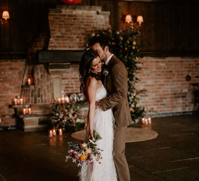 Bride in strapless ivory fringed crepe gown and chapel length veil holding mixed bridal bouquet with garden roses, carnations, peonies and foliage posing with groom in chocolate brown grooms suit in reception room of Willow Marsh Farm Loughborough 