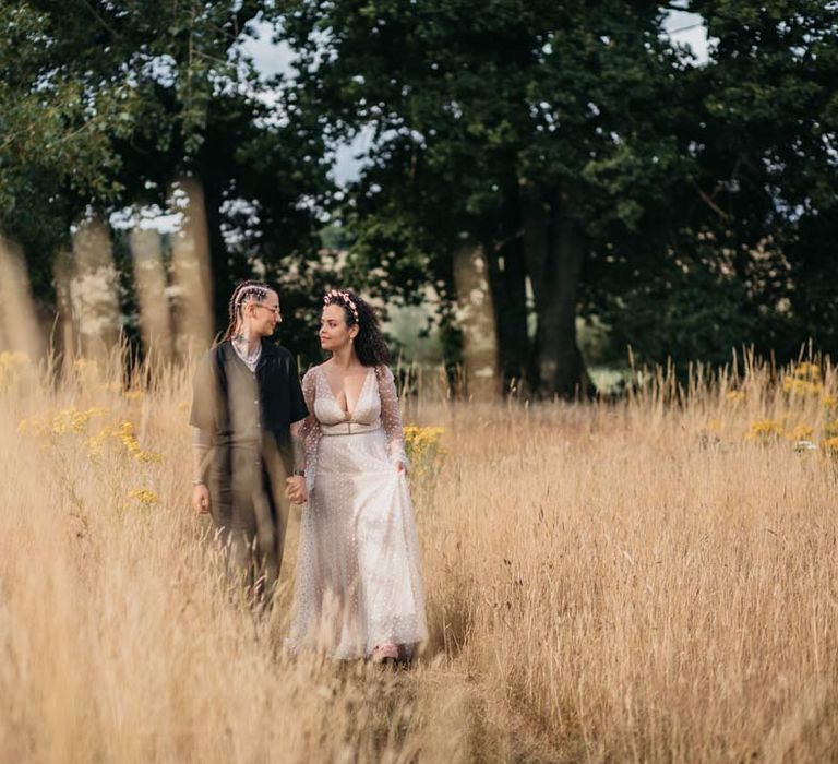 Bride in long sleeve v-neck sheer polka dot overlay sparkly wedding dress and gold celestial bridal headband walking hand in hand with bride in silver chains, glasses, black shirt and black trousers through fields at their at-home Viking themed wedding