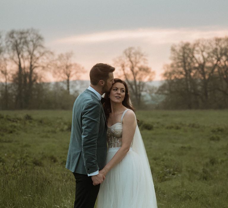 Groom in stunning blue velvet blazer kisses the bride on her forehead at sunset 