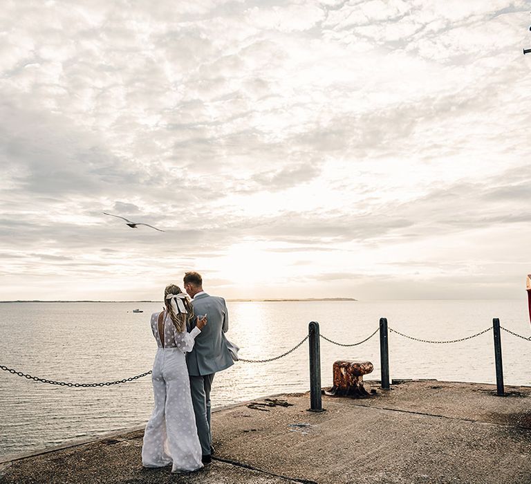 bride in a jumpsuit and groom a grey suit coastal wedding photograph looking at the ocean 