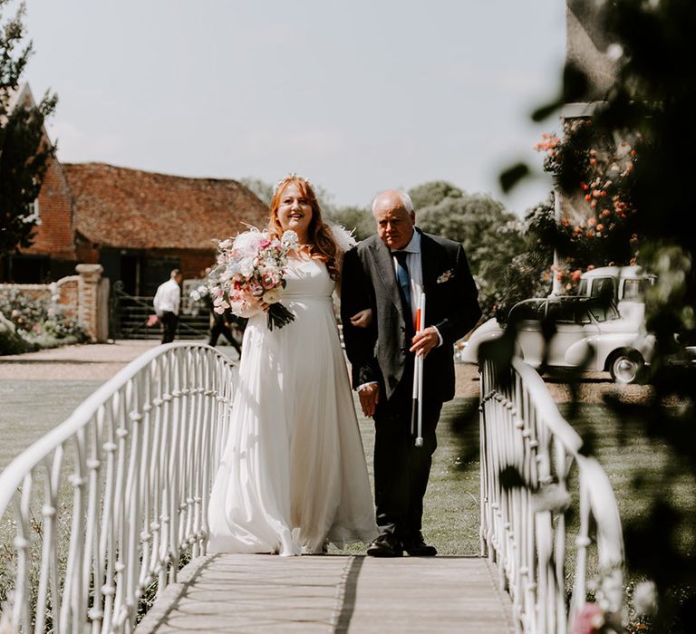 Bride in a leather wedding dress walking with the father of the bride across the bridge to the outdoor ceremony 