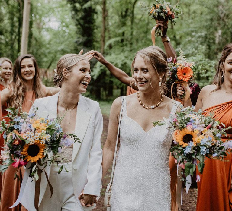 Bride in white wedding suit with lace bodysuit and bride in wedding jumpsuit holding sunflower wedding bouquets 