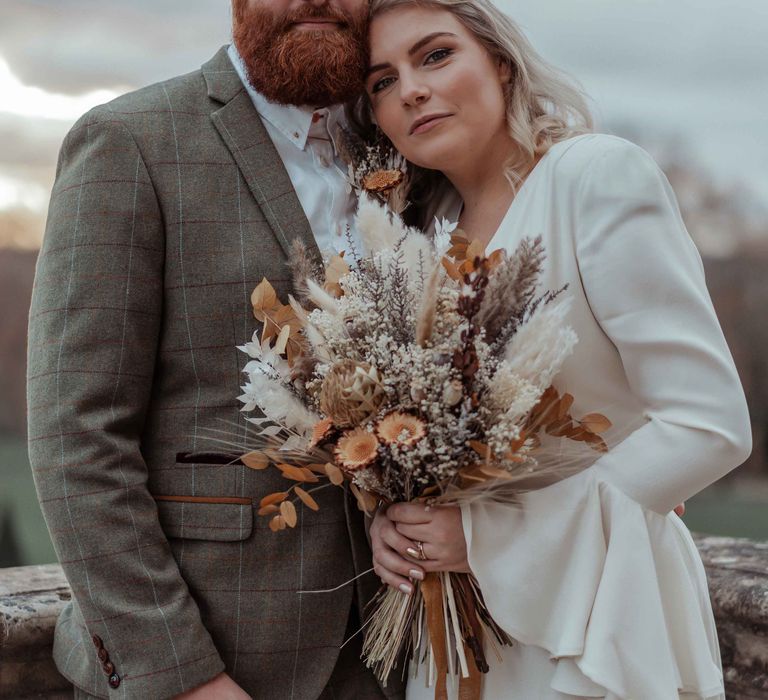 Bride & groom pose featuring tweed suit, Ghost wedding dress and dried floral bouquet 