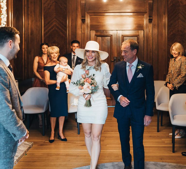 Father of the bride walking his daughter down the aisle with the bride wearing a wedding hat and short Net A Porter Wedding Dress