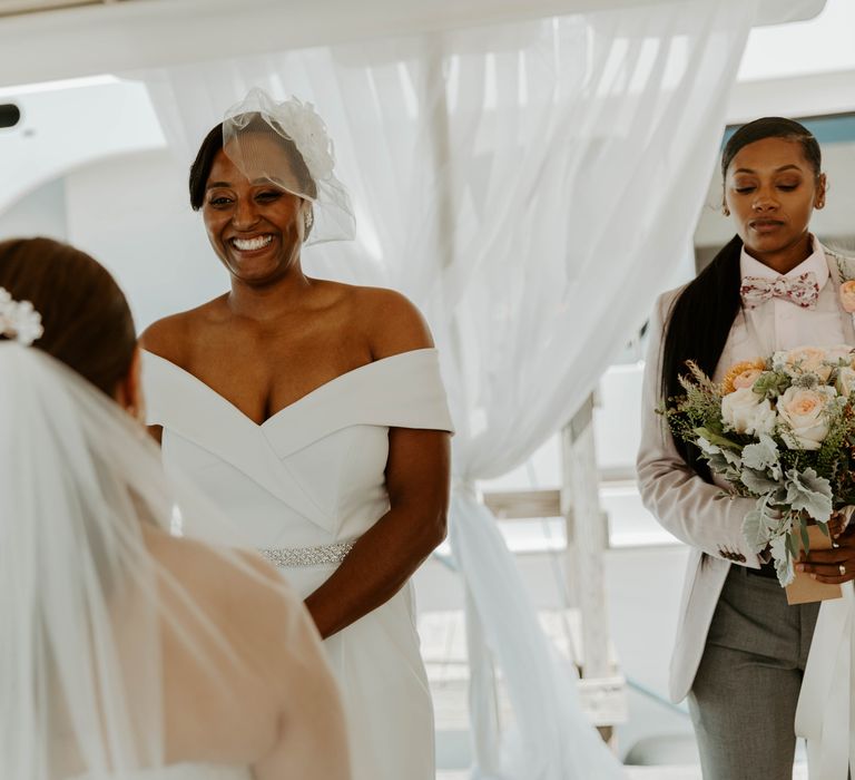 Smiley black bride in a Bardot jumpsuit and blush veil at her same sex wedding ceremony 