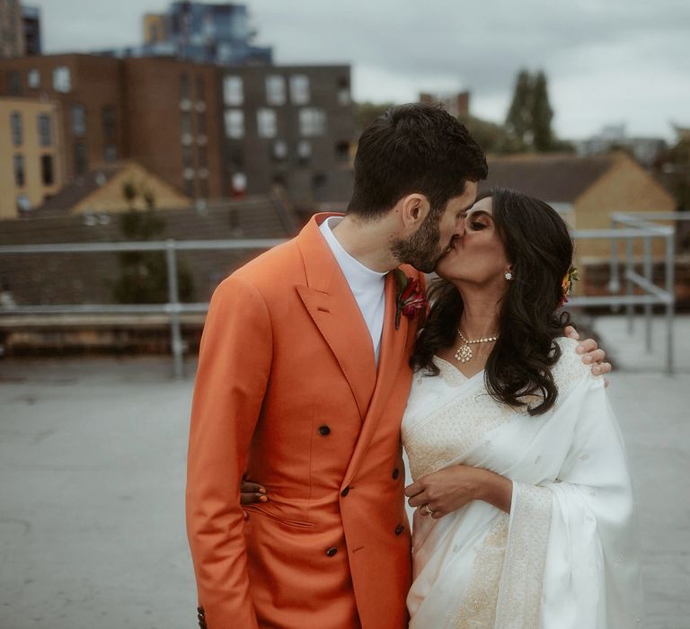 Turkish groom in a white shirt and orange suit kissing his Sri Lankan bride in a white wedding sari 