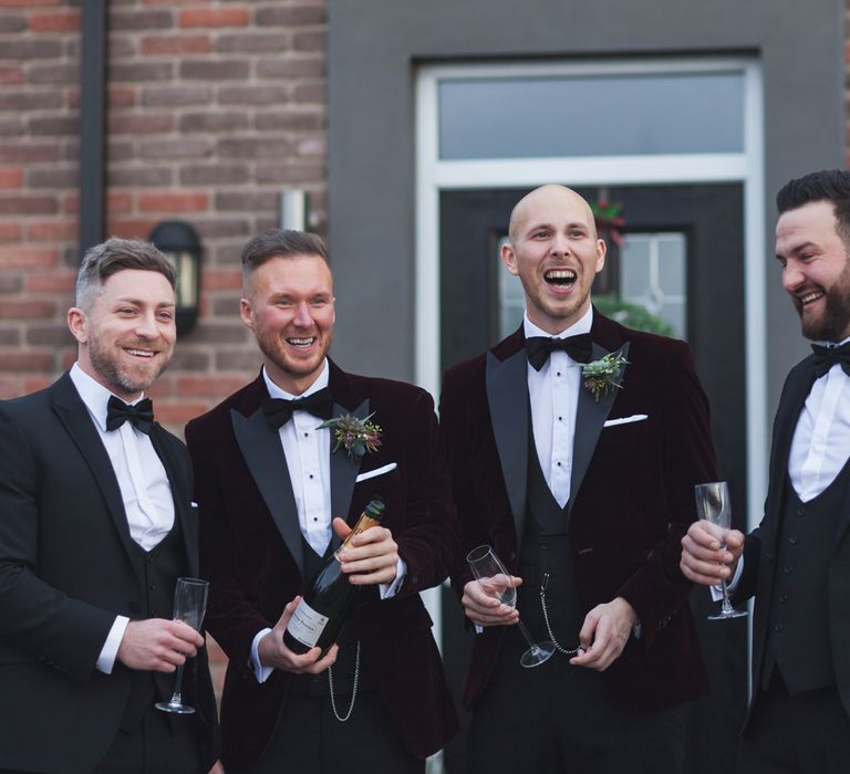 Grooms and groomsmen in burgundy and black tuxedos popping champagne 