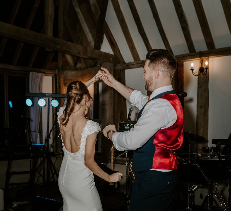 Groom in a navy waistcoat with red silk back twirling his bride in a backless wedding dress with appliqué detail on the dance floor.