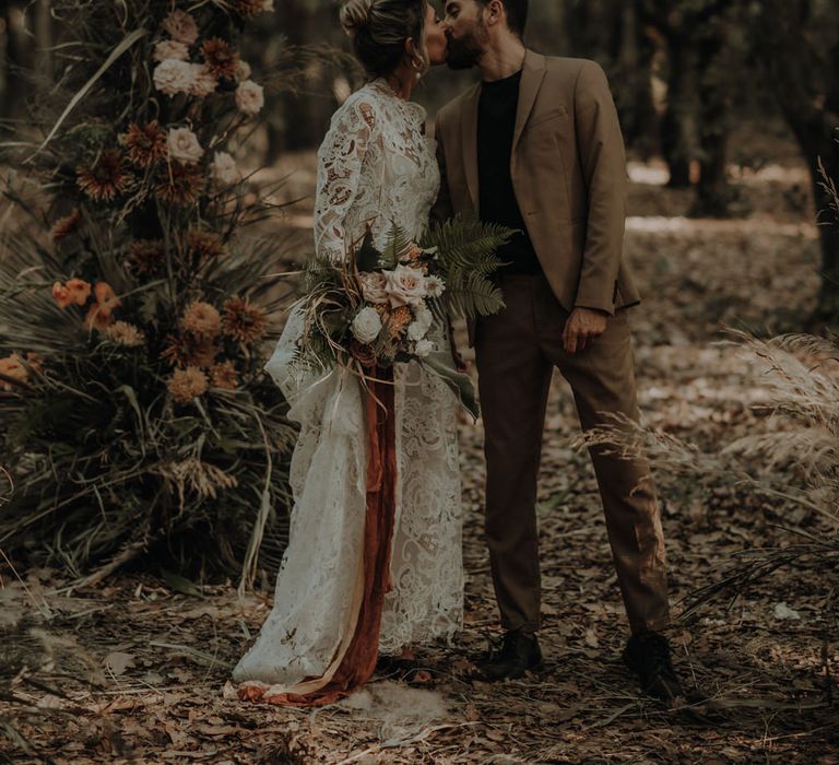 Groom in a beige wedding suit and black t-shirt kissing his bride in a lace wedding dress at their forest elopement 