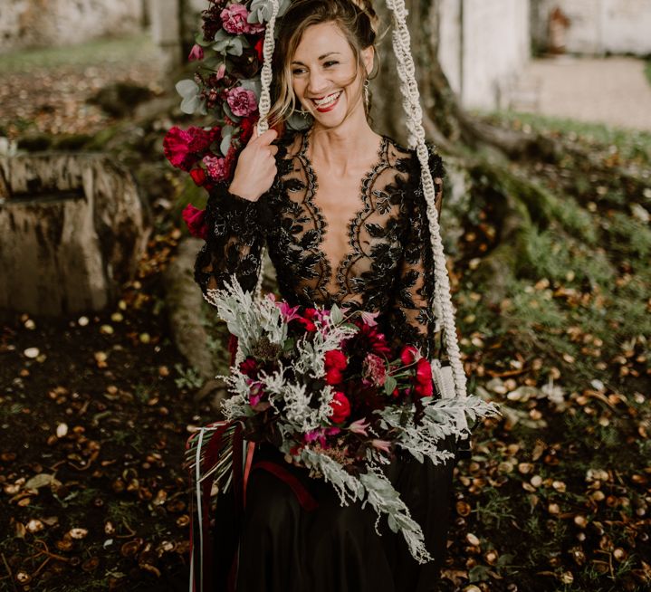 Bride in a black lace wedding dress sitting on a swing decorated with red and green wedding flowers 