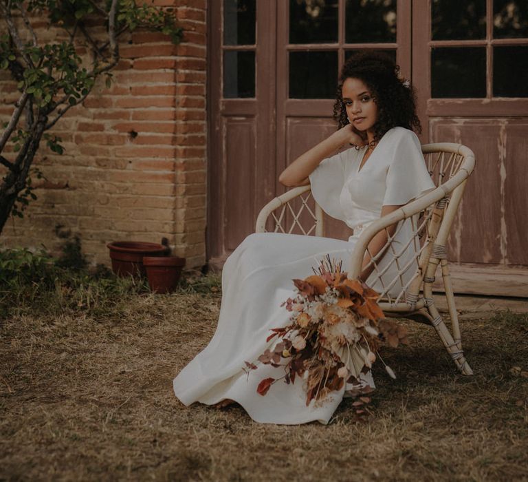 Birde in a minimalist wedding dress with floaty sleeves sitting in a wicker chair holding an orange and brown leaf and flower wedding bouquet 